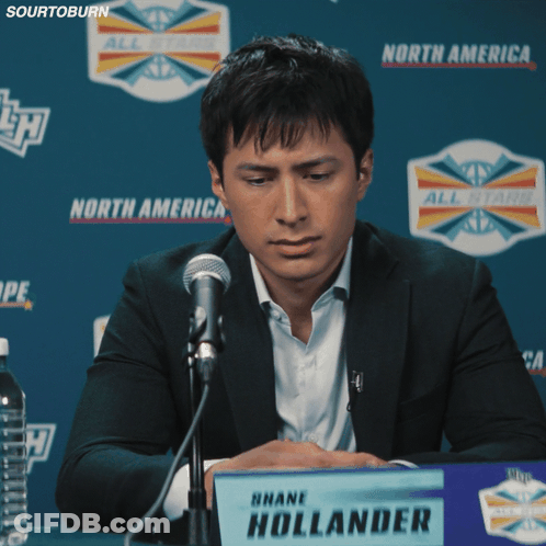 A Man Sitting At A Table With A Sign That Says Shane Hollander On It GIF
