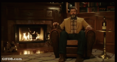 A Man Sitting In A Chair In Front Of A Fireplace With A Bottle Of Wine On A Table GIF