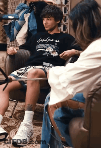 A Man Sitting In A Chair Wearing A Neighborhood Sweatshirt GIF