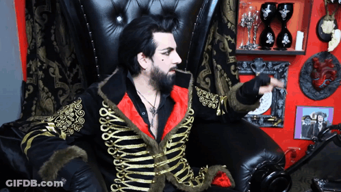 A Man Sitting In A Chair With A Black And Gold Jacket On GIF