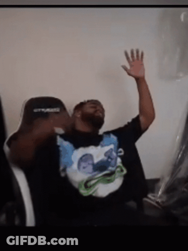 A Man Sitting In A Chair With His Hand In The Air Wearing A Shirt That Says ' Crysis ' On It GIF