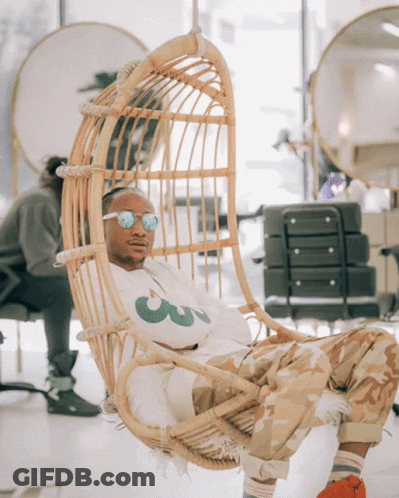 A Man Sitting In A Wicker Chair Wearing Sunglasses And A White Shirt With A Green Leaf On It GIF