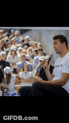A Man Sitting In Front Of A Crowd Holding A Microphone And Wearing A T-Shirt That Says Reein GIF