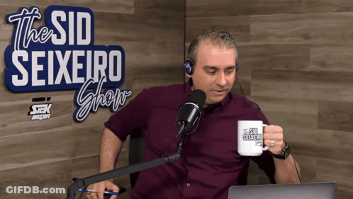 A Man Sitting In Front Of A Microphone With A Mug That Says The Sid Seixeiro Show GIF