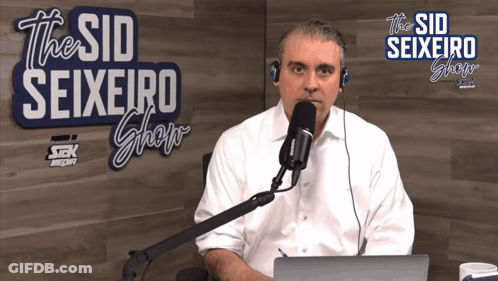 A Man Sitting In Front Of A Microphone With A Sign That Says The Sid Seixeiro Show GIF