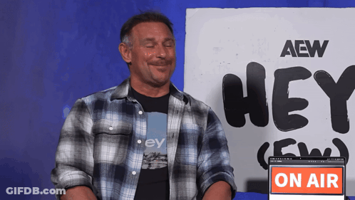 A Man Sitting In Front Of A Sign That Says Hey Aew GIF