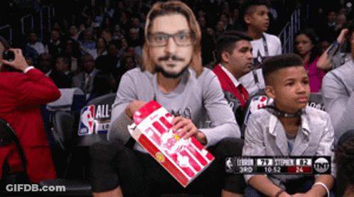 A Man Sitting In The Stands With A Mask On His Face Holding A Bag Of Popcorn GIF