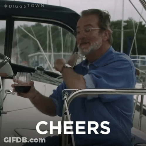A Man Sitting On A Boat Holding A Glass Of Wine With The Word Cheers Behind Him GIF