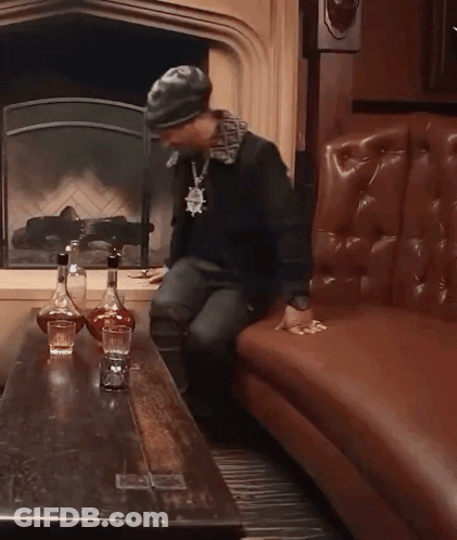 A Man Sitting On A Couch In Front Of A Fireplace With Two Bottles Of Liquor On The Table GIF