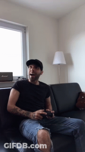 A Man Sitting On A Couch Playing A Video Game With A Marshall Speaker In The Background GIF