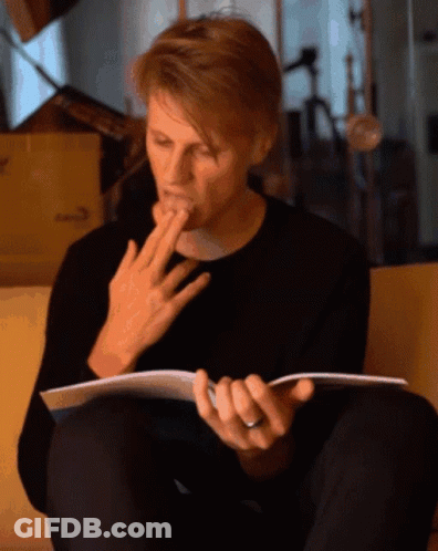 A Man Sitting On A Couch Reading A Book With His Hand On His Mouth GIF