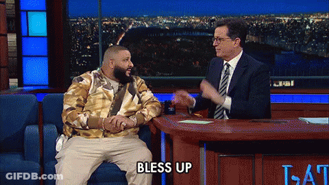 A Man Sitting On A Couch Talking To A Man In A Suit And Tie With The Words Bless Up On The Table GIF