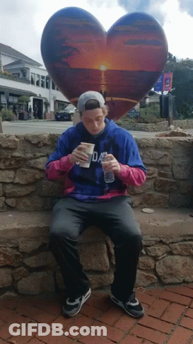 A Man Sitting On A Stone Wall With A Heart Shaped Sculpture In The Background GIF