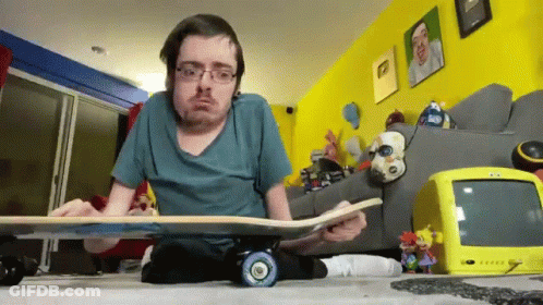 A Man Sitting On The Floor Holding A Skateboard With A Yellow Tv Behind Him GIF