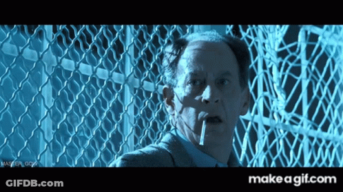 A Man Smoking A Cigarette In Front Of A Chain Link Fence With Make A Gif.com In The Corner GIF