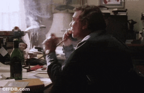 A Man Smoking A Cigarette While Talking On A Phone With A Bottle Of Wine In The Background GIF