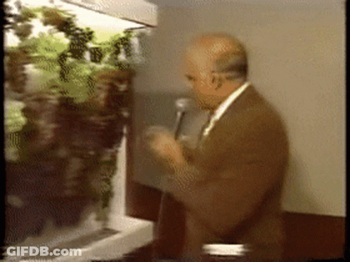 A Man Speaking Into A Microphone In Front Of A Display Of Plants GIF