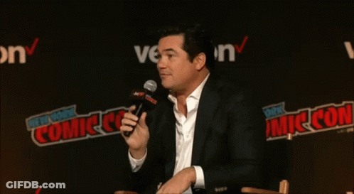 A Man Speaking Into A Microphone In Front Of A Wall That Says New York Comic Con GIF