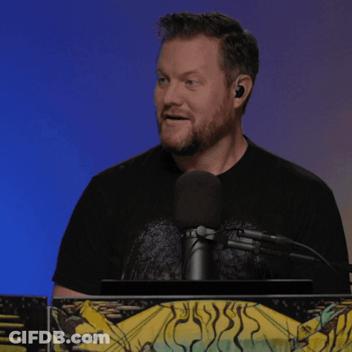 A Man Speaking Into A Microphone While Wearing Earbuds And A Black Shirt GIF