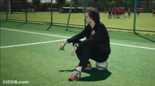 A Man Squatting On A Soccer Ball On A Field With A Team In The Background GIF