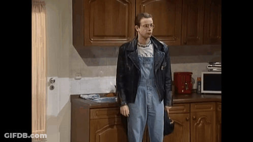 A Man Standing In A Kitchen Wearing Overalls And A Leather Jacket GIF