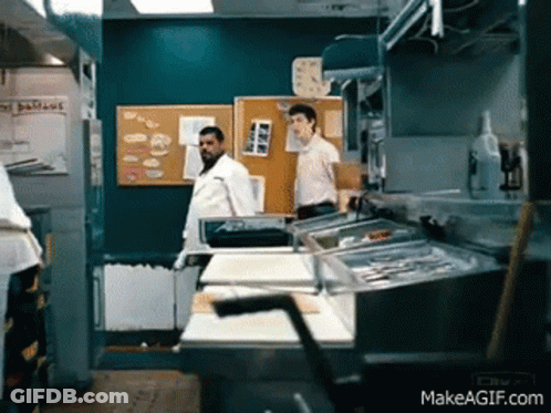 A Man Standing In A Kitchen With A Sign On The Wall That Says Delicious GIF