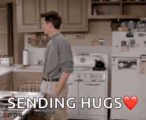 A Man Standing In A Kitchen With The Words " Sending Hugs " Above Him GIF