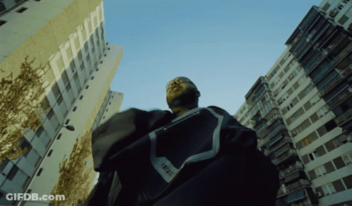 A Man Standing In Front Of A Building With The Word Trust On His Shirt GIF