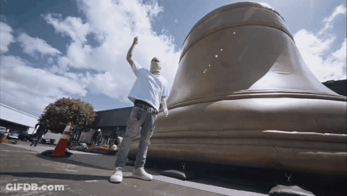 A Man Standing In Front Of A Large Bell With His Hand In The Air GIF
