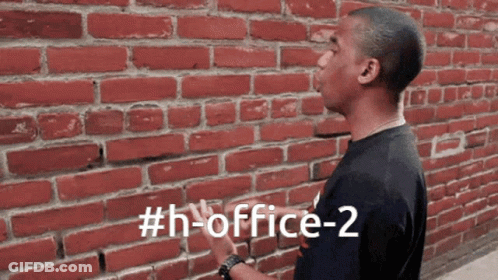 A Man Standing In Front Of A Red Brick Wall With The Hashtag # H-Office-2 GIF