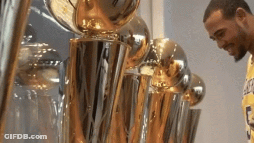 A Man Standing In Front Of A Row Of Trophies Including One That Says Lakers On It GIF