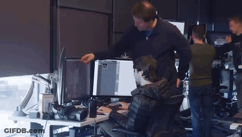 A Man Standing Next To A Man Sitting At A Desk Looking At A Computer Screen GIF