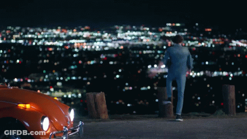 A Man Standing Next To An Orange Car Looking At A City GIF