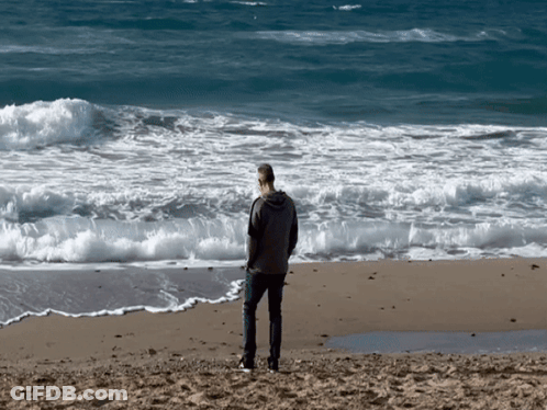 A Man Standing On A Beach Looking At The Waves GIF