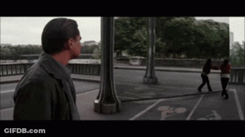 A Man Standing On A Bridge Looking At A Woman Walking On The Sidewalk GIF
