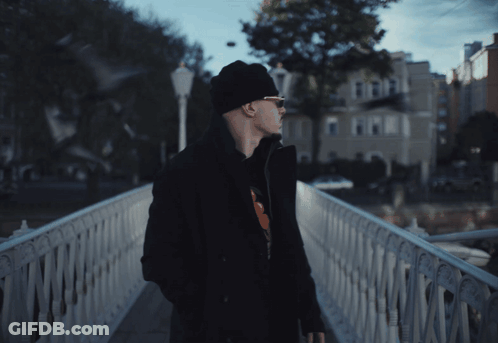 A Man Standing On A Bridge With A Building In The Background GIF