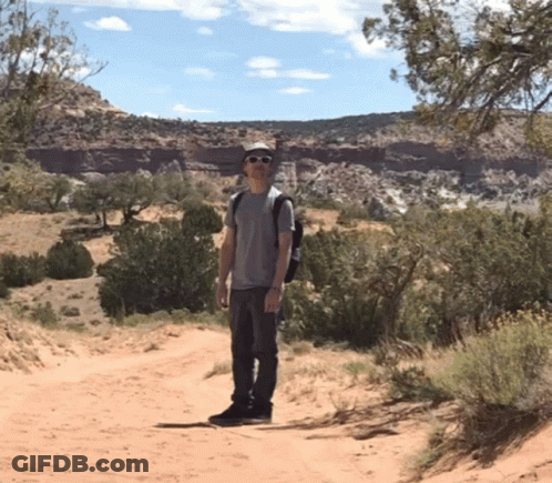 A Man Standing On A Dirt Road Wearing Sunglasses And A Hat GIF