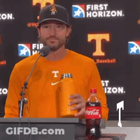 A Man Stands At A Podium Holding A Coca Cola Bottle And A Water Bottle GIF