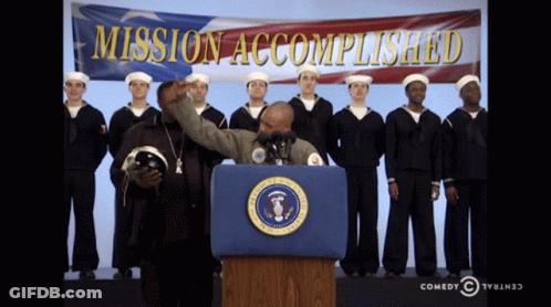 A Man Stands At A Podium In Front Of A Mission Accomplished Banner GIF