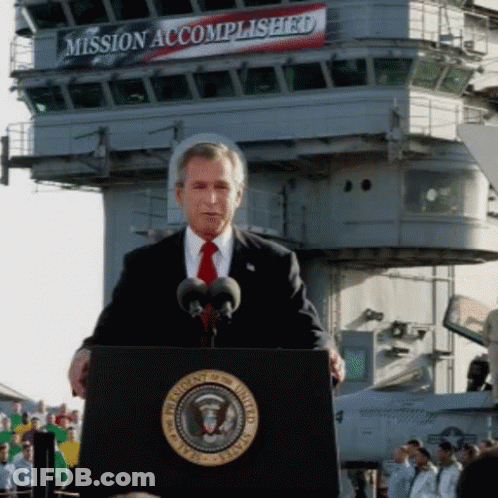 A Man Stands At A Podium In Front Of A Sign That Says Mission Accomplished On It GIF