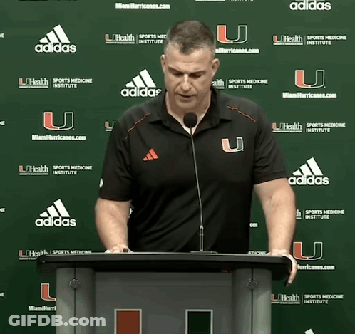 A Man Stands At A Podium In Front Of A Wall With Adidas Logos GIF