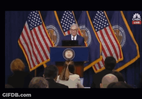 A Man Stands At A Podium In Front Of American Flags With Cnbc Live On The Screen Behind Him GIF
