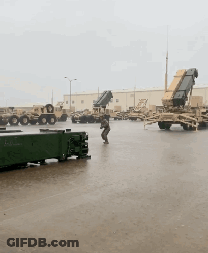 A Man Stands In A Parking Lot With Military Vehicles And A Container That Says ' Army ' On It GIF
