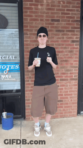 A Man Stands In Front Of A Brick Building Holding A Cup Of Coffee GIF