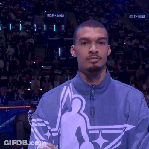 A Man Stands In Front Of A Crowd Wearing A Blue Jacket With A Basketball Player On It GIF