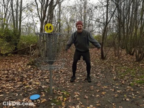 A Man Stands In Front Of A Frisbee Basket With A Yellow Sign That Says 14 GIF