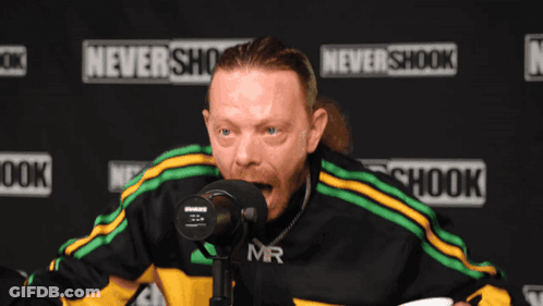 A Man Stands In Front Of A Microphone In Front Of A Sign That Says Never Shook GIF