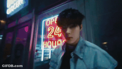A Man Stands In Front Of A Neon Sign That Says 24 Hours GIF