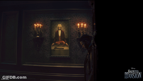 A Man Stands In Front Of A Painting Of A Man In A Tuxedo With Candles In A Haunted Mansion Room GIF