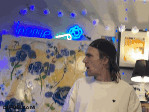A Man Stands In Front Of A Painting With A Neon Sign That Says Home Behind Him GIF